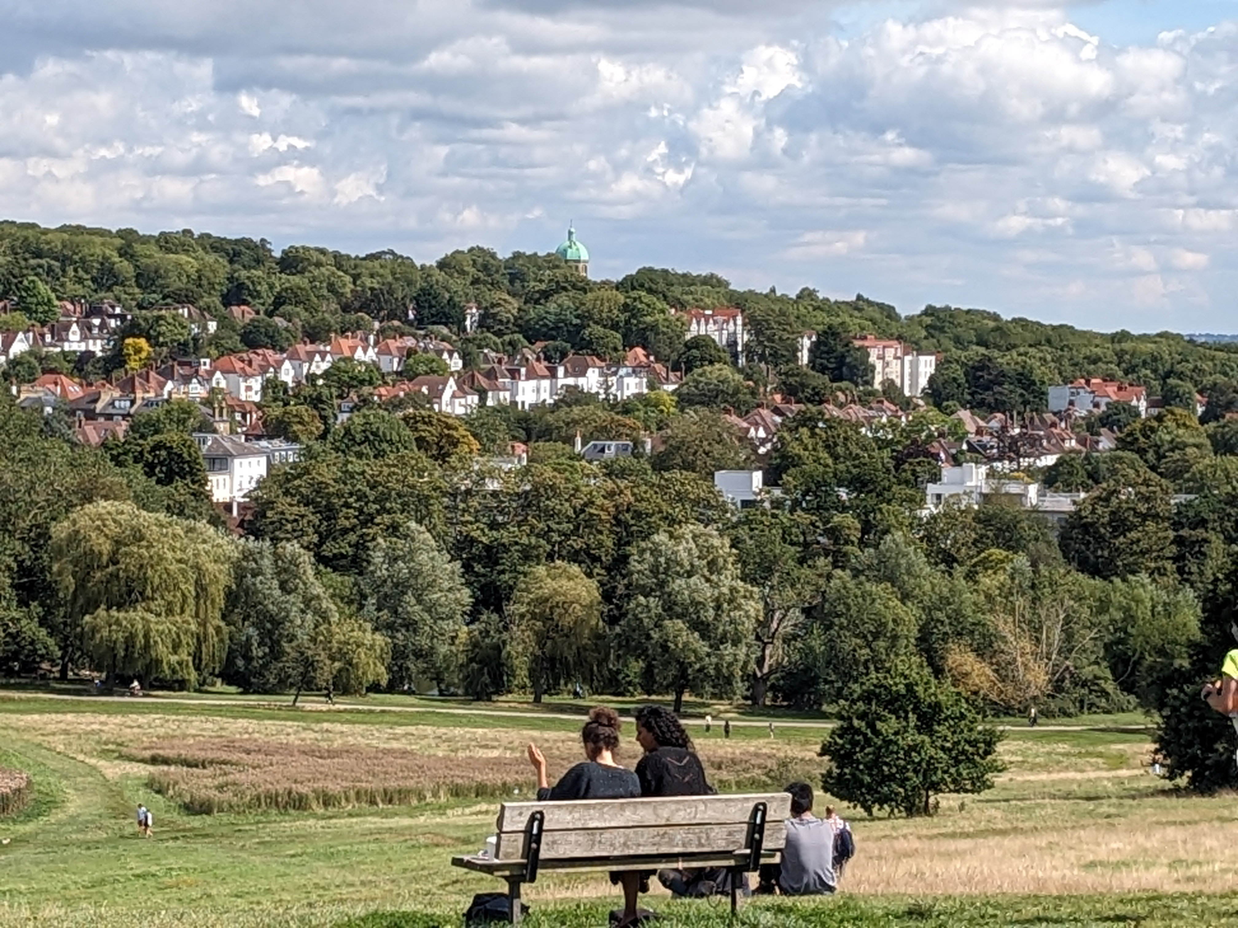 a hillside with houses among clusters of trees. two people sit on a park bench in the foreground.